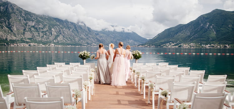 Bride and bridesmaids in front of lake and mountains with wedding chairs framing image